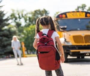 A little girl wearing a backpack and standing in front of a bus.